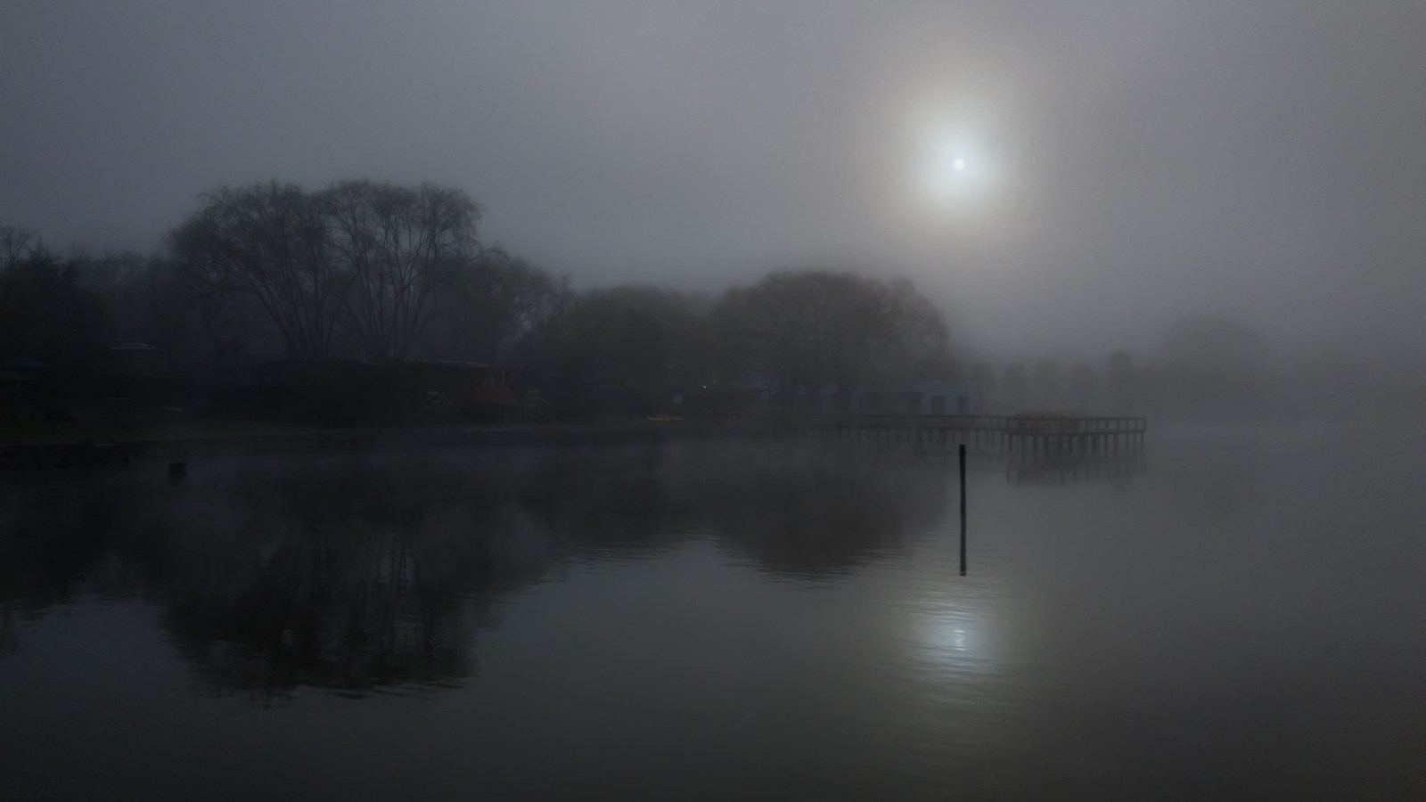 Neblina de la mañana sobre el lago, con el sol detrás y el muelle a la derecha